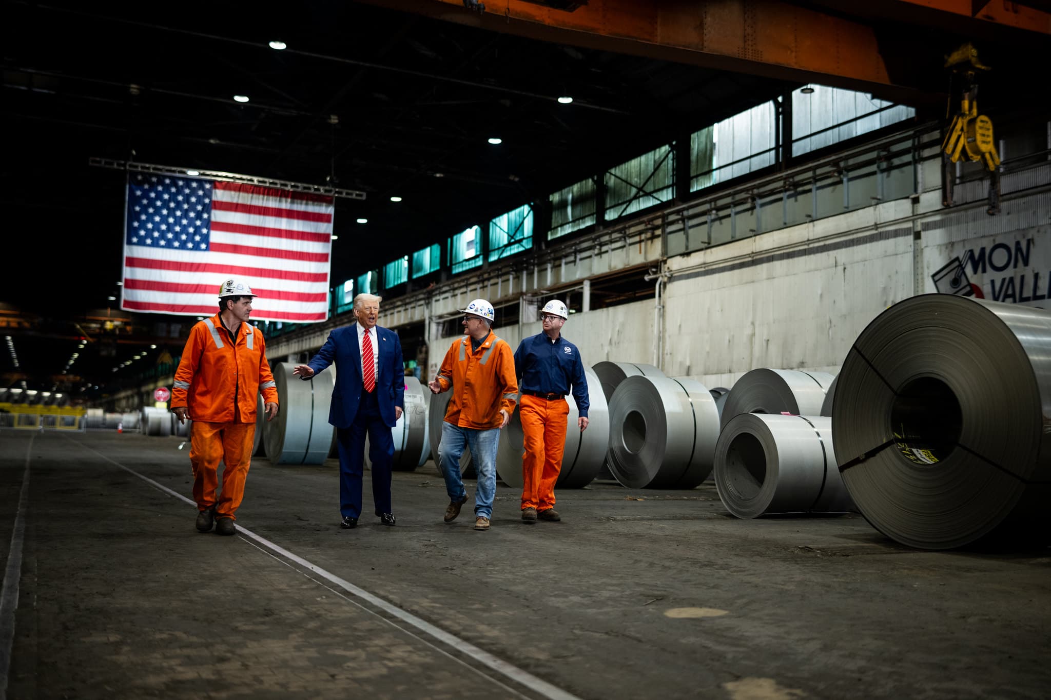 President Donald J. Trump walking through a U.S. Steel mill with workers, American flag in background.