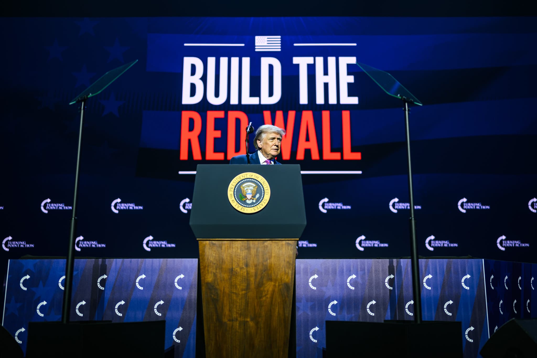 President Trump speaking at Turning Point Action in front of a 'Build the Red Wall' backdrop.